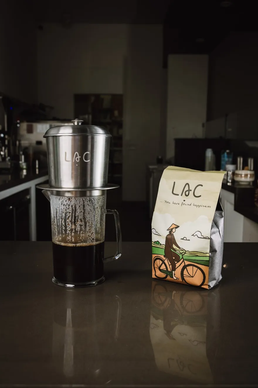 Phin filter dripping Vietnamese coffee into a pitcher beside a bag of Lạc Coffee beans
